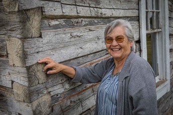 Lilyrose Meyers, a respected knowledge holder, next to a rustic log cabin belonging to her ancestors in Metis Crossing, Alberta.