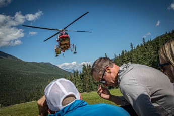 The Bell helicopter hovers barely three metres above the alpine grass, whilst those waiting crouch down, their hands covering their faces. Behind them, spruce trees tower upwards.