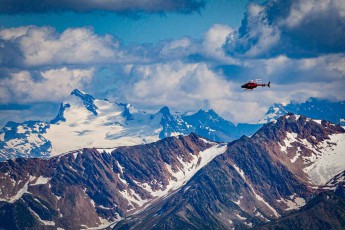 A carmine-red helicopter crosses the jagged ridgelines of the Cariboo Mountains, firn fields and scree slopes falling away beneath it in shades of grey.
