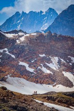 Hikers cross a strip of old snow below North Canoe Glacier – small figures against ochre rock and the serrated peaks of the Cariboos.