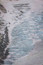 Blue-and-white glacial ice breaks up into sharp seracs, separated by strips of snow that look like seams. Moraine debris flanks the slope – an abstract work of art.