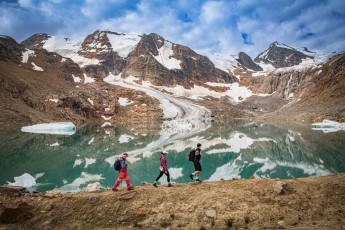 Three hikers trace the moraine path above Buehler Lake. Still, turquoise water mirrors glacier and jagged ridge — loose ice floes slow the eye before it reaches the peaks.