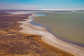 Die salzverkrustete Uferlinie schneidet zwischen ockerfarbenem Festland und milchigem Türkis. Mineralien färben das seichte Wasser in Schichten von Schlamm bis Aquamarin.