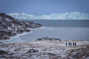 Fünf Gestalten auf dem Holzweg unterhalb des Ilulissat Icefjord Centre, dahinter die Kante des Eisfjords – der Weg führt hinunter zur zugefrorenen Diskobucht.
