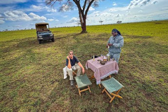 Kaffeepause mitten in der Ebene, im Schatten einer Akazie: Klapphocker, Tischtuch, Kanne und Blechbecher – Guide Hesekiel Msangi schenkt ein, Annette wartet.