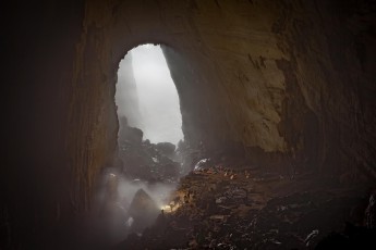 Son Doong Cave, Vietnam: Blick auf Camp 1, unser Nachtlager. Im Vordergrund leuchtet ein Begleiter in Richtung einer Wolkenschwade. Im Hintergrund fällt der Lichtschein von der Öffnung der Doline 300 Meter tief auf den Boden. Dolinen sind natürliche Schächte, die durch Einsturz der Höhlendecke entstehen.
