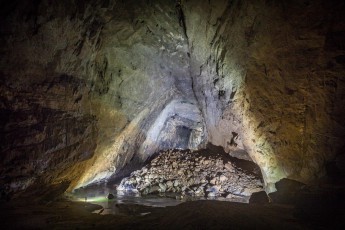 Blick durch den Mittelraum der Hang En Cave. Hier mischt sich das Licht unserer Höhlenlampen mit dem Tageslicht, welches durch den Ausgang der Höhle hereinströmt. Schon die Hang En ist riesig: Oben auf dem Schottenhaufen erkennt beim genauen Hinsehen das grüne Trikot eines Expeditionsmitglieds vor einem dunklen Quadrat im Hintergrund.