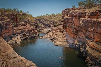 In der Bell Gorge haben sich zwei Schwimmer eine natürliche Oase erobert – das kristallklare Wasser sammelt sich zwischen den schroffen Sandsteinwänden zu einem perfekten Naturpool, während ringsum die in Millionen von Jahren geschliffenen Felsformationen in warmen Ockertönen in der Kimberley-Sonne glühen.
