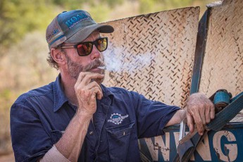 Abschleppwagenfahrer Darren bei einer wohlverdienten Zigarettenpause nn seinem Fahrzeug auf der Gibb River Road. Hinter verspiegelten Sonnengläsern und einem wettergebräunten Gesicht verbirgt sich jemand, der schon unzählige gestrandete Reisende aus misslichen Lagen befreit hat. Seine sonnengegerbte Haut und die entspannte Haltung erzählen Geschichten von endlosen Kilometern auf Schotterpisten und Begegnungen mit den Launen der Kimberley-Wildnis.