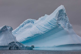 Frozen waves trace across the flank of this iceberg – marks of ancient melt and freeze cycles. The deep blue waterline marks the zone of constant erosion.