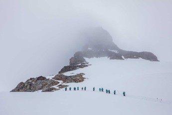 In single file, a group trudges across the snowfield at Meusnier Point. Fog swallows the summit, only dark rock outcrops pierce the white.