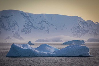 Like sculptures, icebergs rest before the mighty mountain backdrop of Antarctica. The yellowish sky heralds the low sun that sets close to midnight.