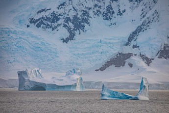 Two icebergs in different stages of weathering drift in front of the mighty glacier landscape, each iceberg telling its own story of pressure, time and decay. On the right, the bright blue reveals ancient ice that was once hidden.